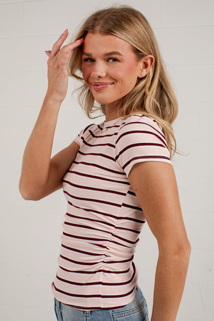 A woman wearing a fitted short-sleeve t-shirt with horizontal burgundy stripes on a light pink base, paired with blue jeans, posing against a white brick wall.