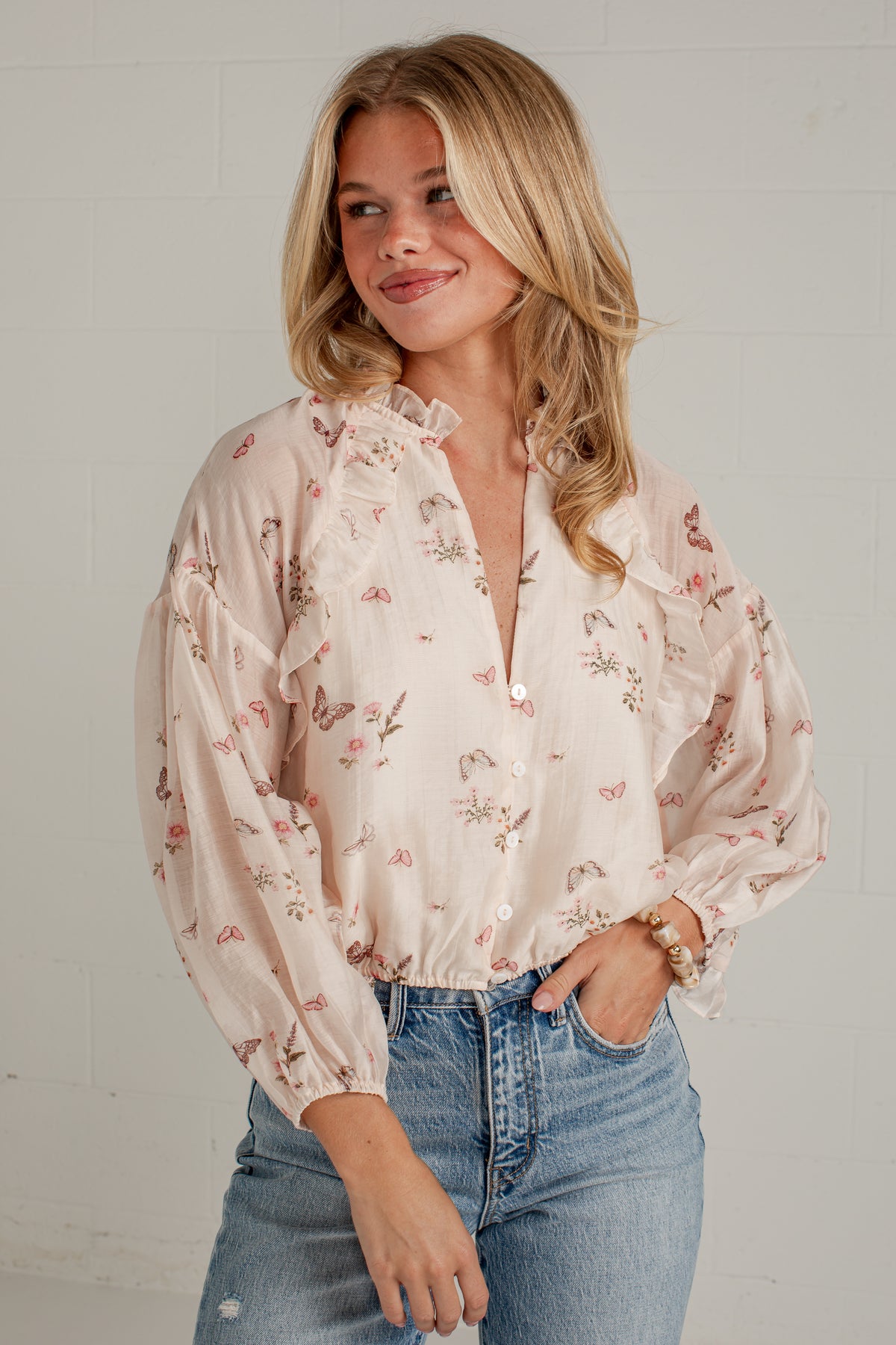 A woman wearing a light beige long-sleeve blouse with a delicate pink and green floral print, ruffled collar, and puff sleeves, paired with blue jeans against a white brick wall.