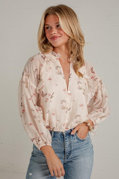 A woman wearing a light beige long-sleeve blouse with a delicate pink and green floral print, ruffled collar, and puff sleeves, paired with blue jeans against a white brick wall.
