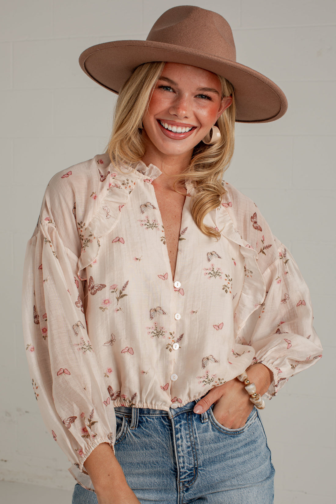 A woman wearing a light beige floral long-sleeve blouse, blue jeans, and a brown wide-brim hat, posing against a light gray wall.