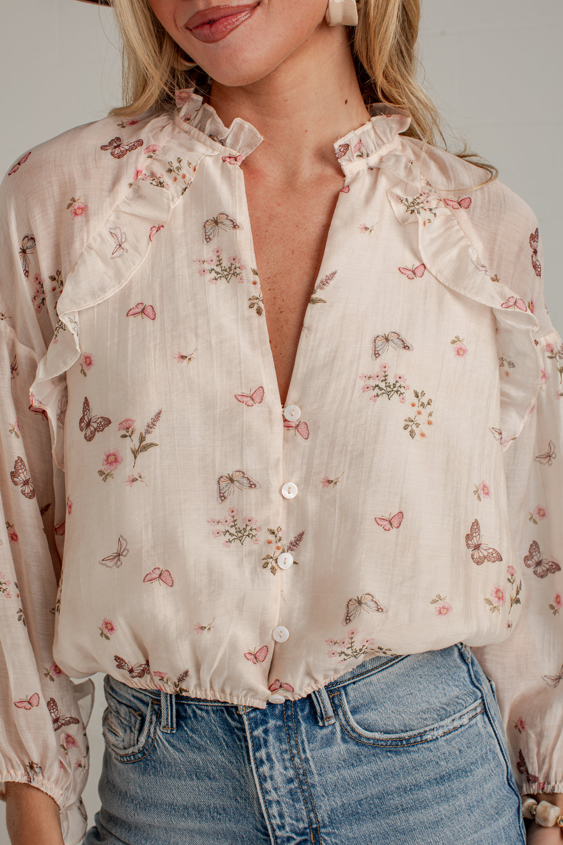 A close-up of a woman wearing a cream-colored long-sleeve blouse with a delicate pink and green butterfly and floral print, paired with blue jeans.