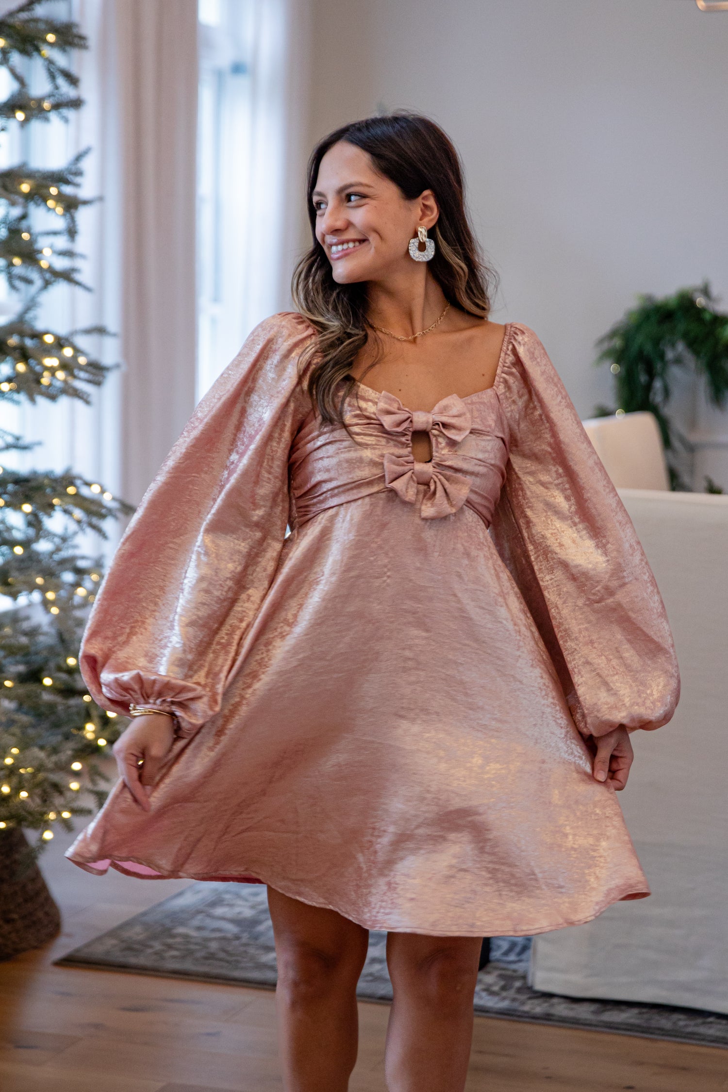 Woman wearing a pink dress with bow detail in a room with a Christmas tree.