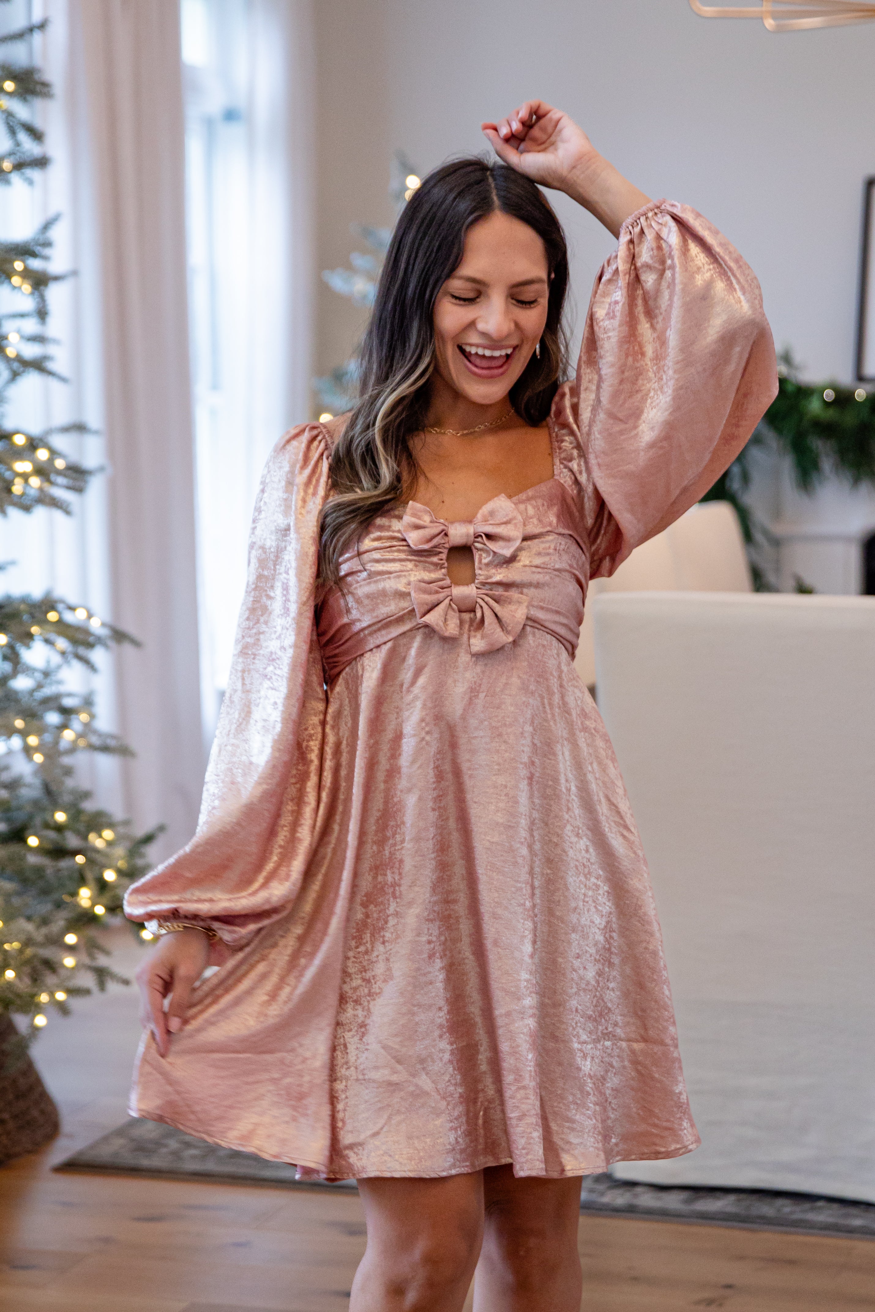 Woman in a pink dress with a bow standing in a living room with Christmas decorations.