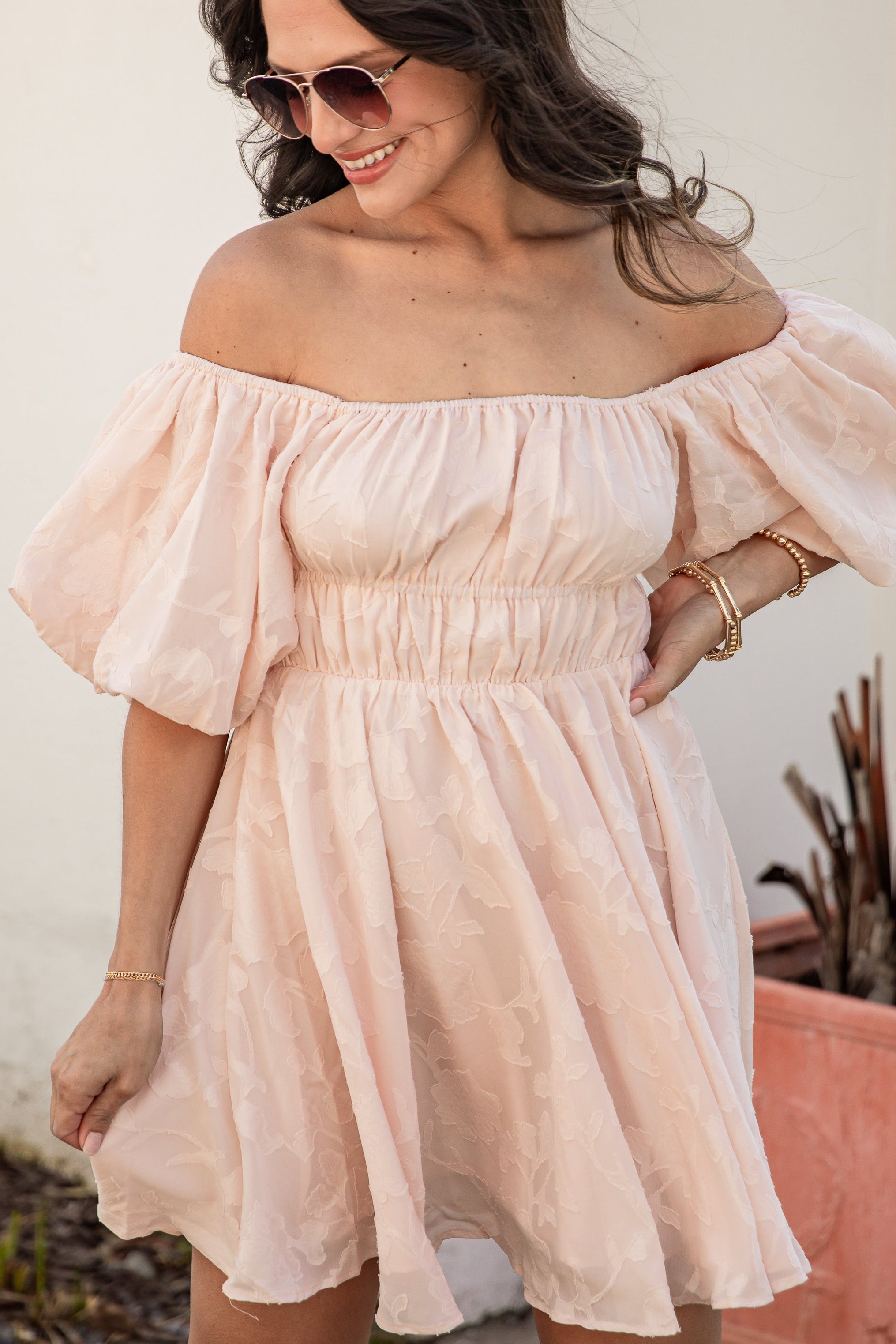 Woman wearing a light pink off-shoulder dress standing against a white wall.