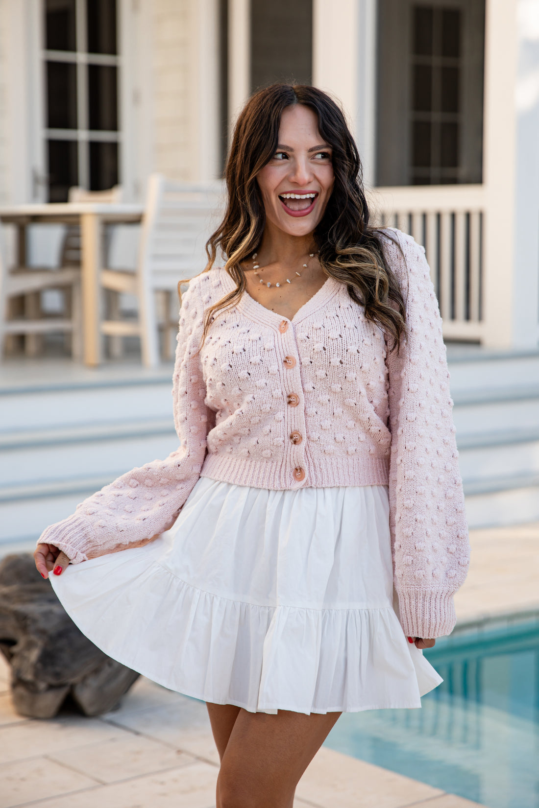 Woman wearing a pink cardigan and white skirt standing by a pool.