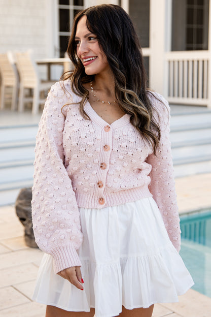 Woman wearing a pink cardigan over a white dress by a poolside.