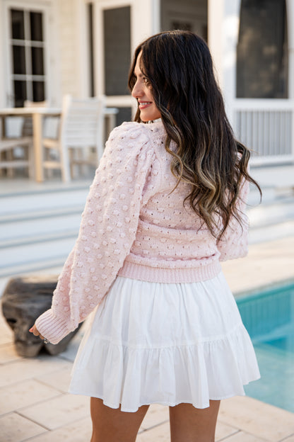 Woman in a pink sweater and white skirt standing by a poolside.