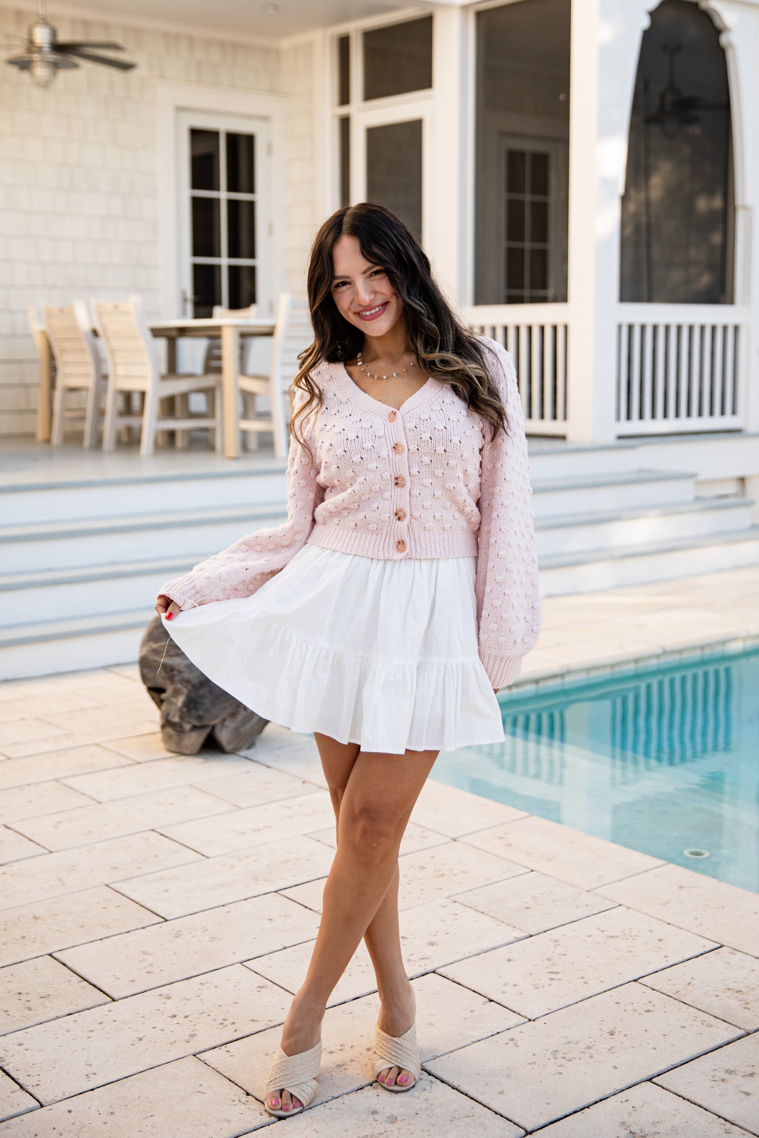 Woman in a pink cardigan and white skirt standing by a poolside.