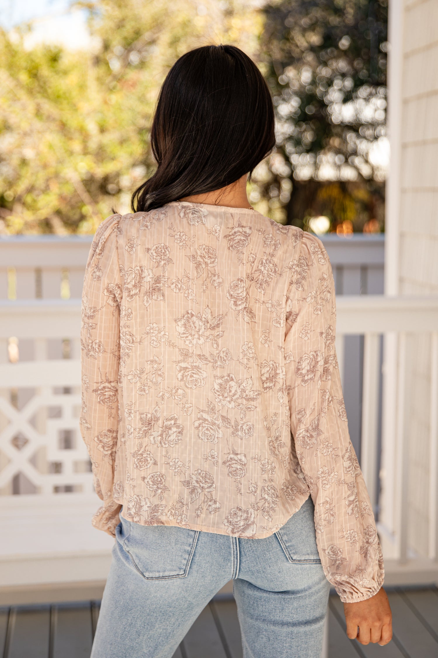 Woman wearing a beige floral blouse and light blue jeans on a porch.