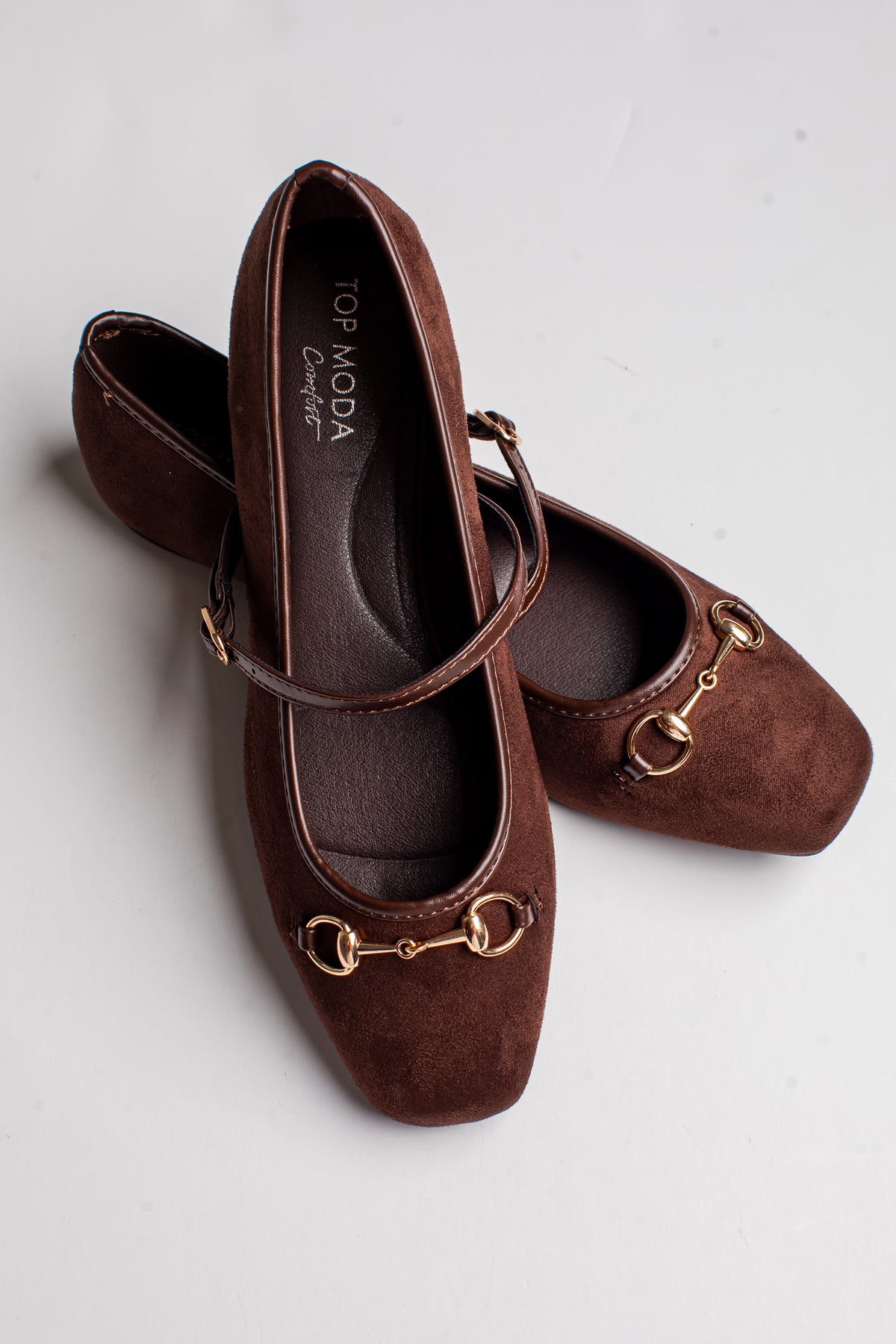 A pair of dark brown suede Mary Jane flats with gold-tone horsebit hardware and a thin strap, displayed on a light gray background. The brand name &