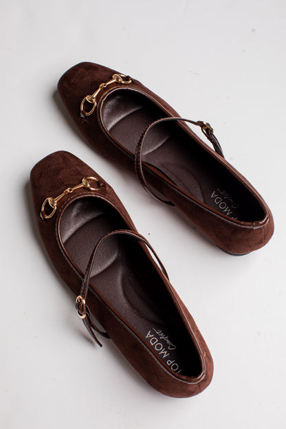 A pair of brown suede flat shoes with gold-tone horsebit hardware and thin ankle straps, displayed on a light gray background. The brand name &