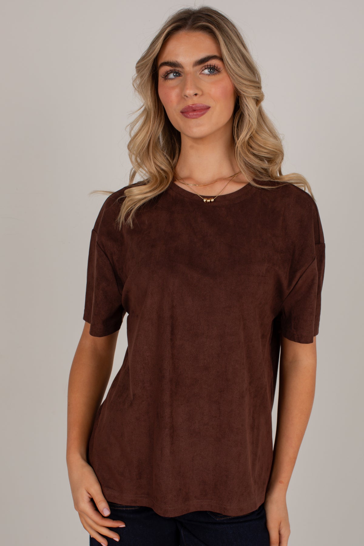 A woman wearing a short-sleeve crewneck top in a dark brown suede-like fabric, photographed against a plain light gray studio background.