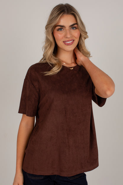 A woman wearing a brown short-sleeve top with a soft suede-like texture, posed against a solid light gray background.