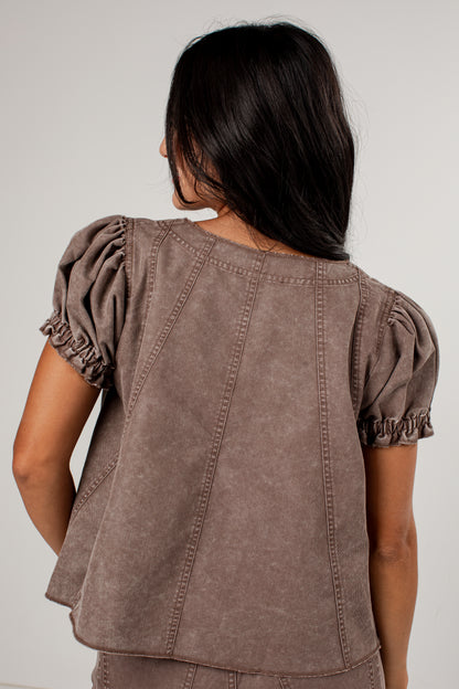A woman viewed from the back wearing a cropped taupe-brown top with puffed short sleeves and vertical seam detailing, against a plain light gray studio background.