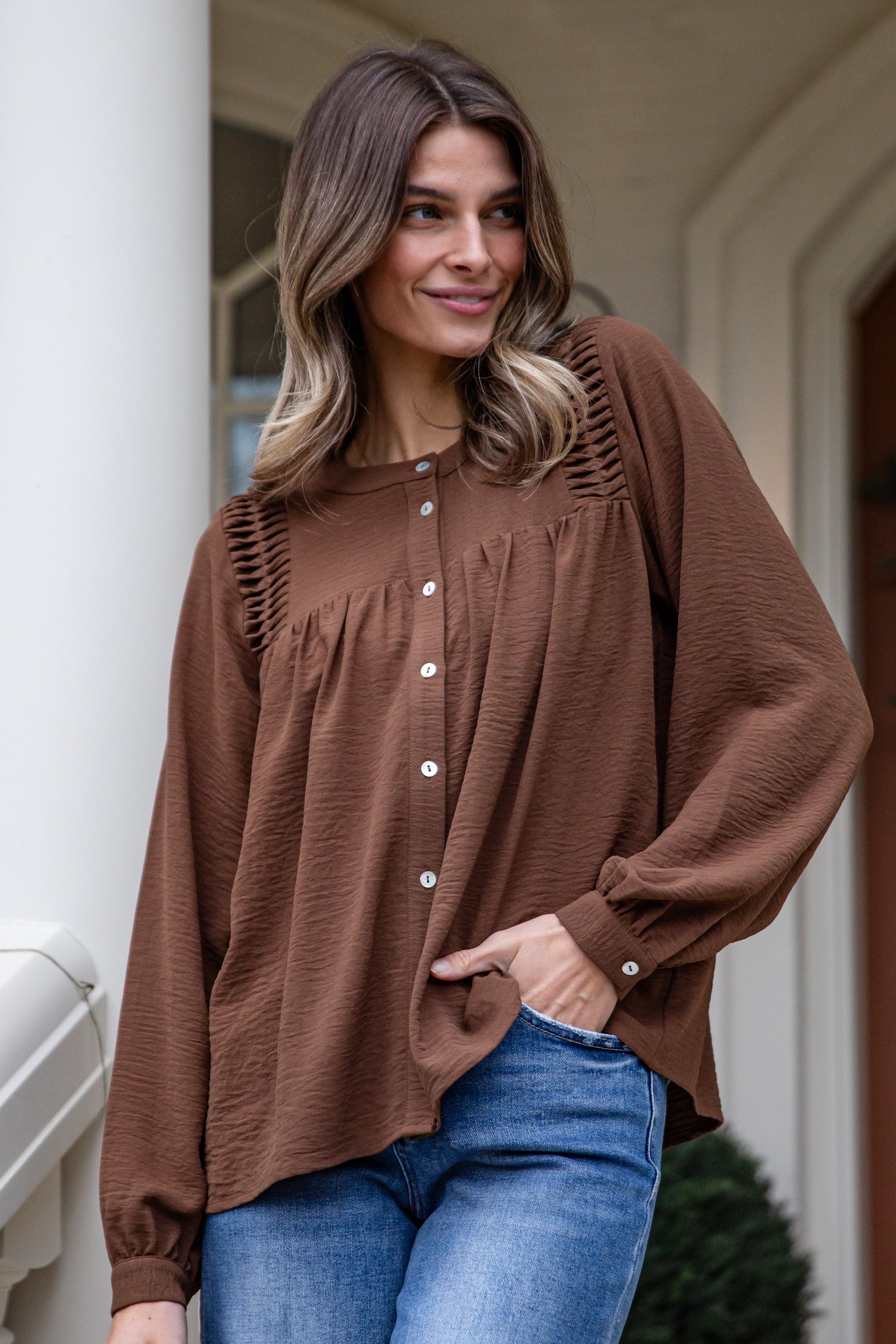 Woman wearing a brown blouse and blue jeans standing on a porch.