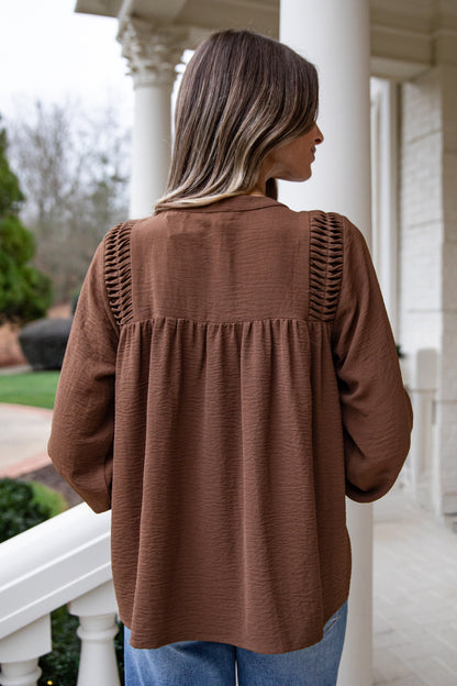 Woman wearing a brown blouse with intricate shoulder designs, standing on a porch.