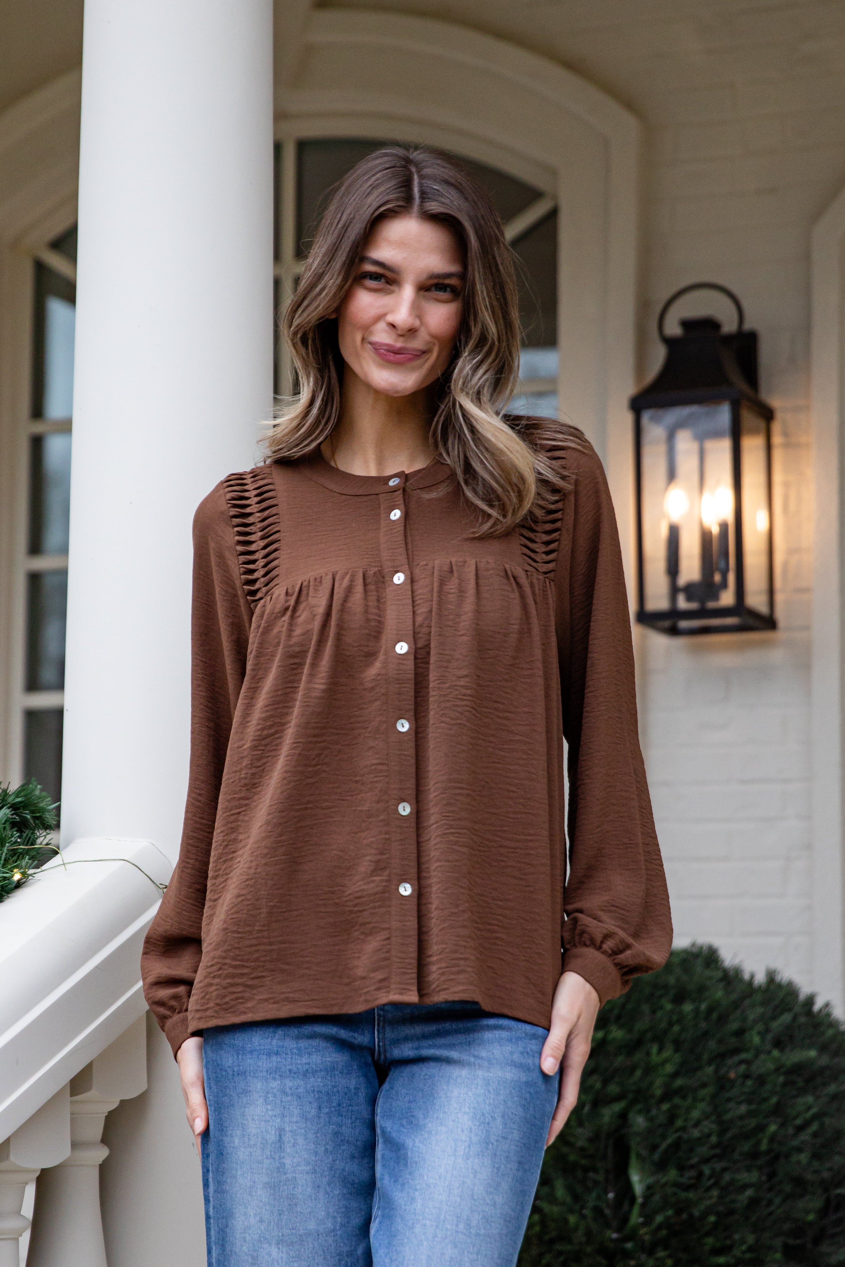 Woman wearing a brown blouse and blue jeans standing on a porch.