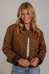 A smiling blonde woman wearing a brown utility-style jacket with dark brown corduroy collar and cuffs, layered over a white top and blue jeans, standing against a light gray wall.