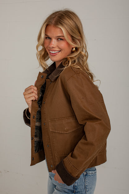 A woman wearing a brown waxed cotton utility jacket with dark brown corduroy collar and cuffs, paired with blue jeans, posing against a light gray brick wall.