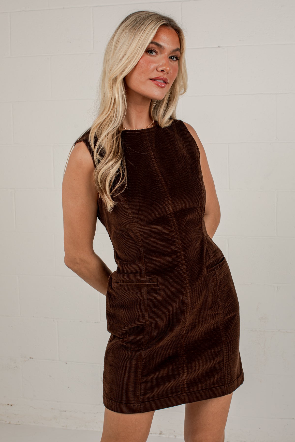 A blonde woman wearing a sleeveless dark brown corduroy dress, standing against a white painted brick wall.
