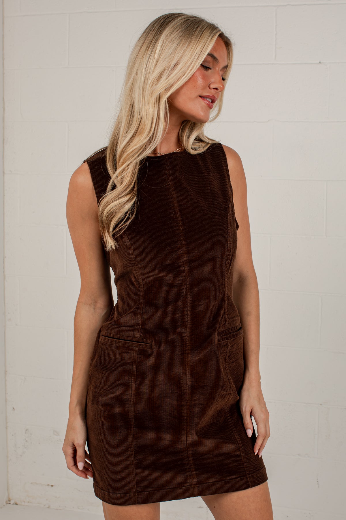 A blonde woman wearing a sleeveless brown corduroy dress with front patch pockets, standing against a white painted brick wall.