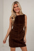 A blonde woman wearing a sleeveless brown corduroy mini dress with front patch pockets, standing against a white brick wall.