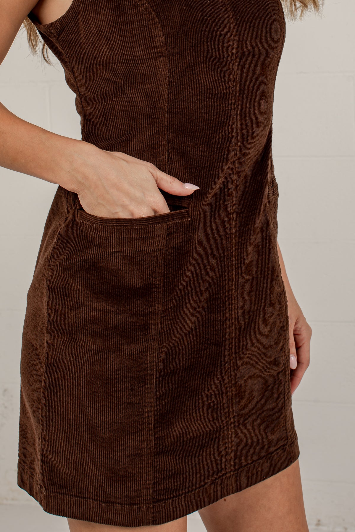 A close-up of a woman wearing a brown corduroy dress with a front patch pocket, shown from the shoulders to mid-thigh against a light neutral background.