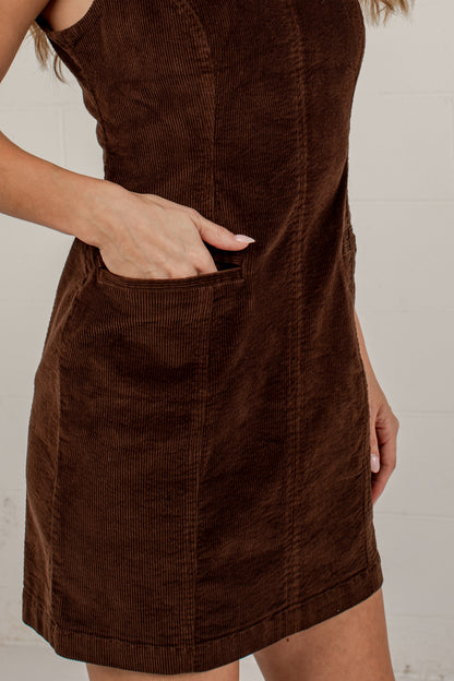 A close-up of a woman wearing a brown corduroy dress with a front patch pocket, shown from the shoulders to mid-thigh against a light neutral background.