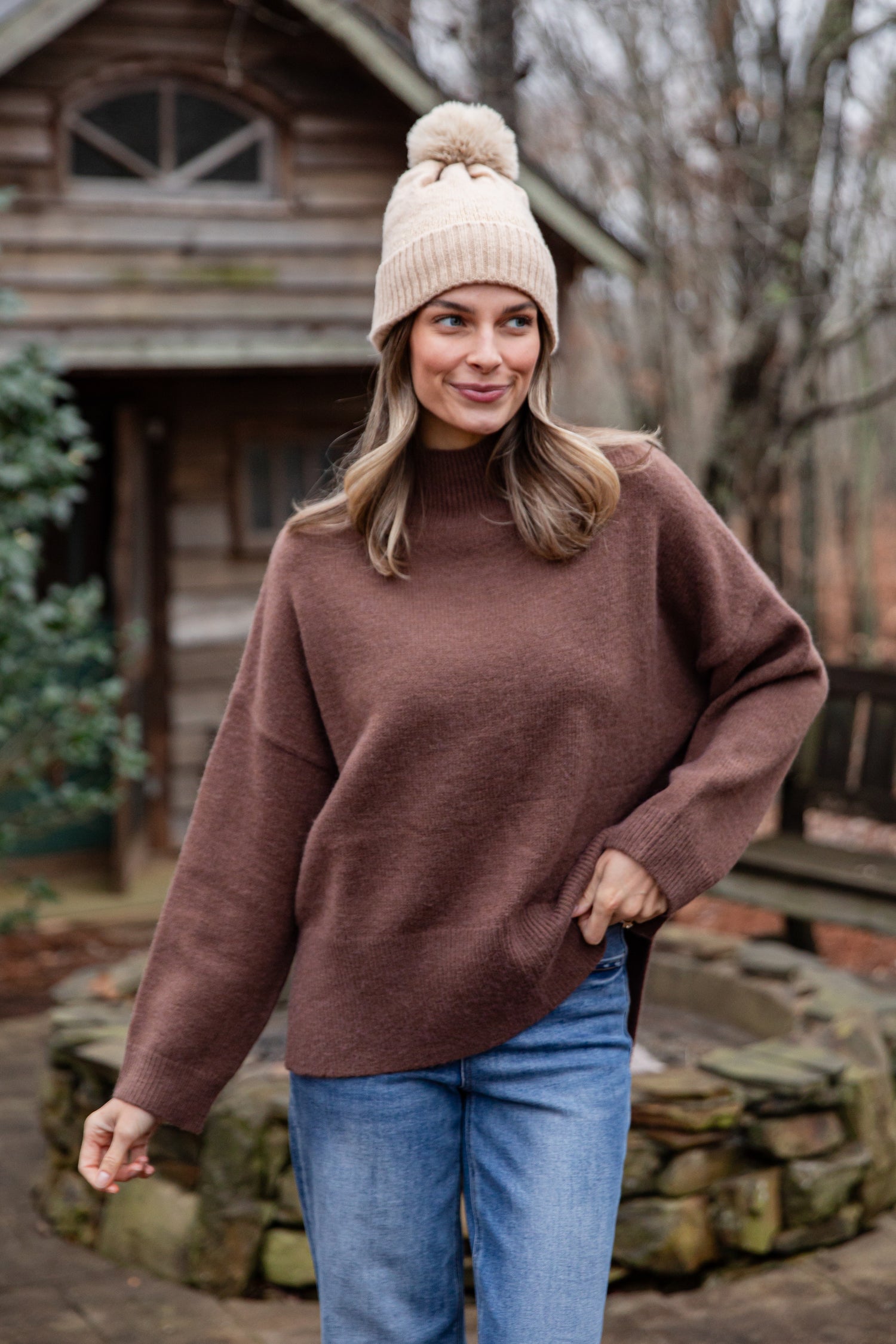 Woman wearing a brown sweater and beige beanie outdoors near a wooden cabin.