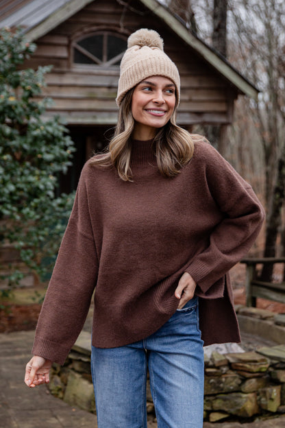 Woman wearing a brown sweater and beige beanie standing in front of a wooden cabin.