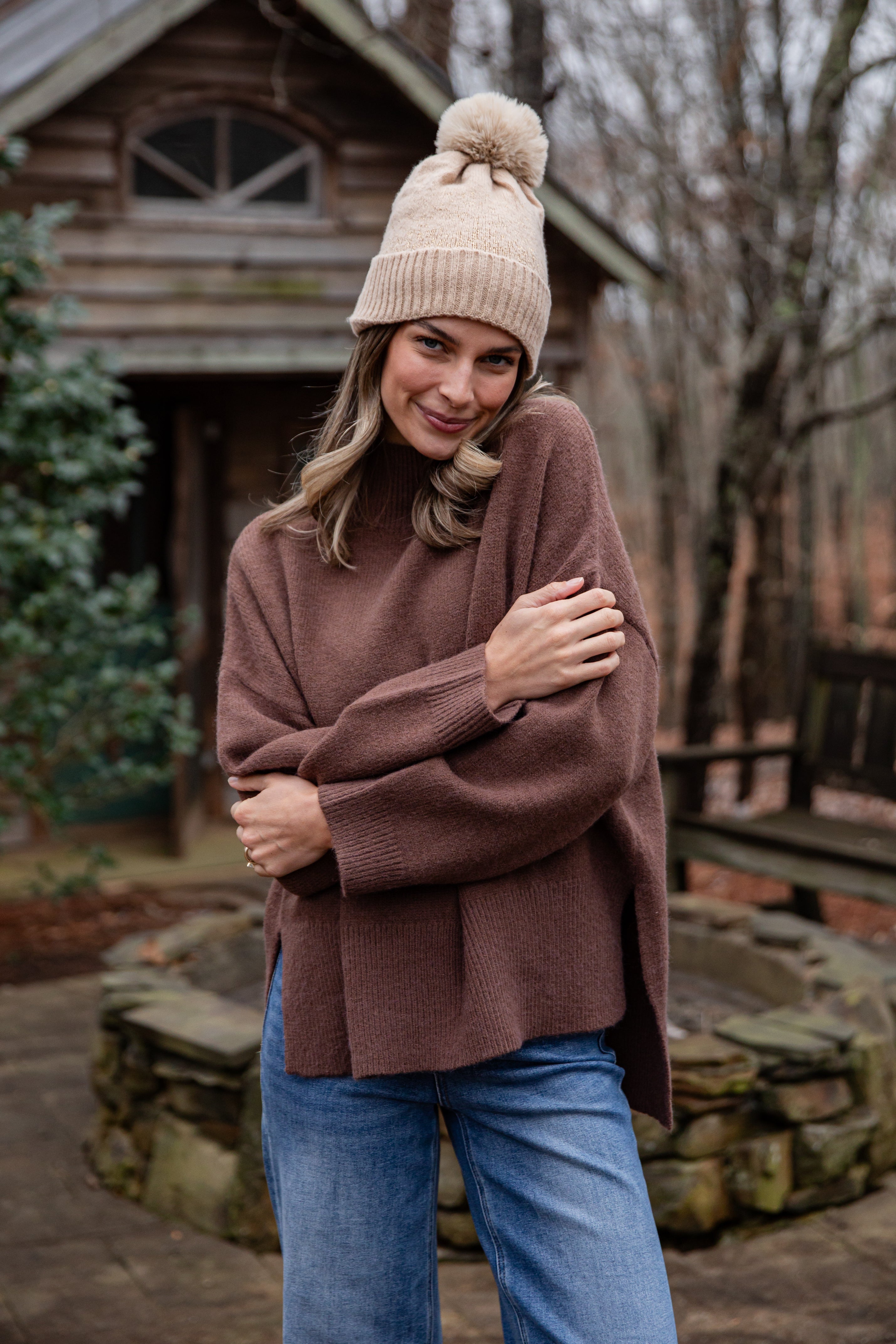 Woman wearing a brown sweater and beige knit hat standing in front of a wooden cabin.