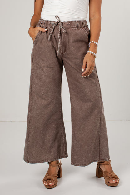 A person wearing wide-leg cropped pants in a washed taupe-brown color, styled with a white top and brown block-heel sandals against a light gray studio background.