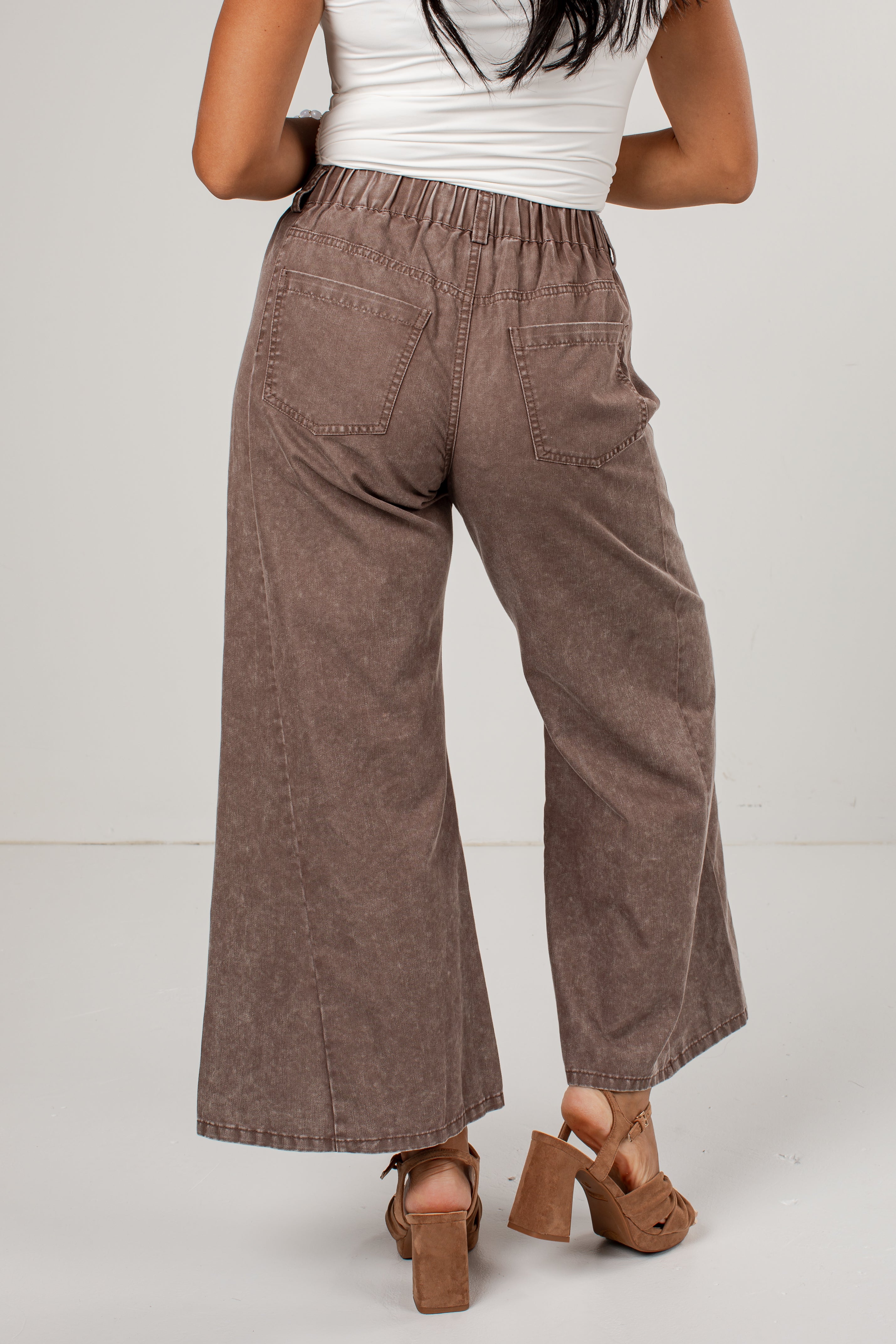 A person shown from the back wearing wide-leg cropped pants in a washed taupe-brown color, paired with a white top and brown platform sandals, against a light gray studio background.