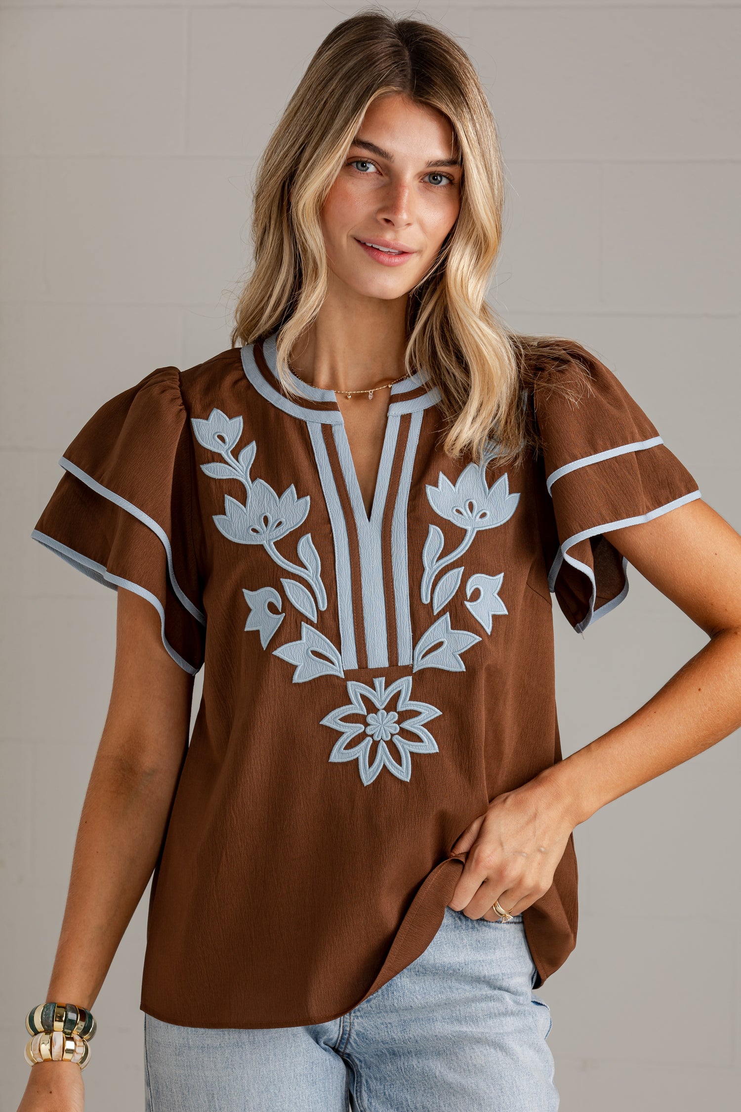 A woman wearing a brown short-sleeve blouse with white floral embroidery and striped trim, paired with light blue jeans, against a neutral gray background.
