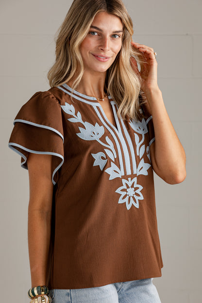 A woman wearing a brown short-sleeve top with white floral embroidery on the front, paired with light blue jeans, photographed against a neutral light gray background.