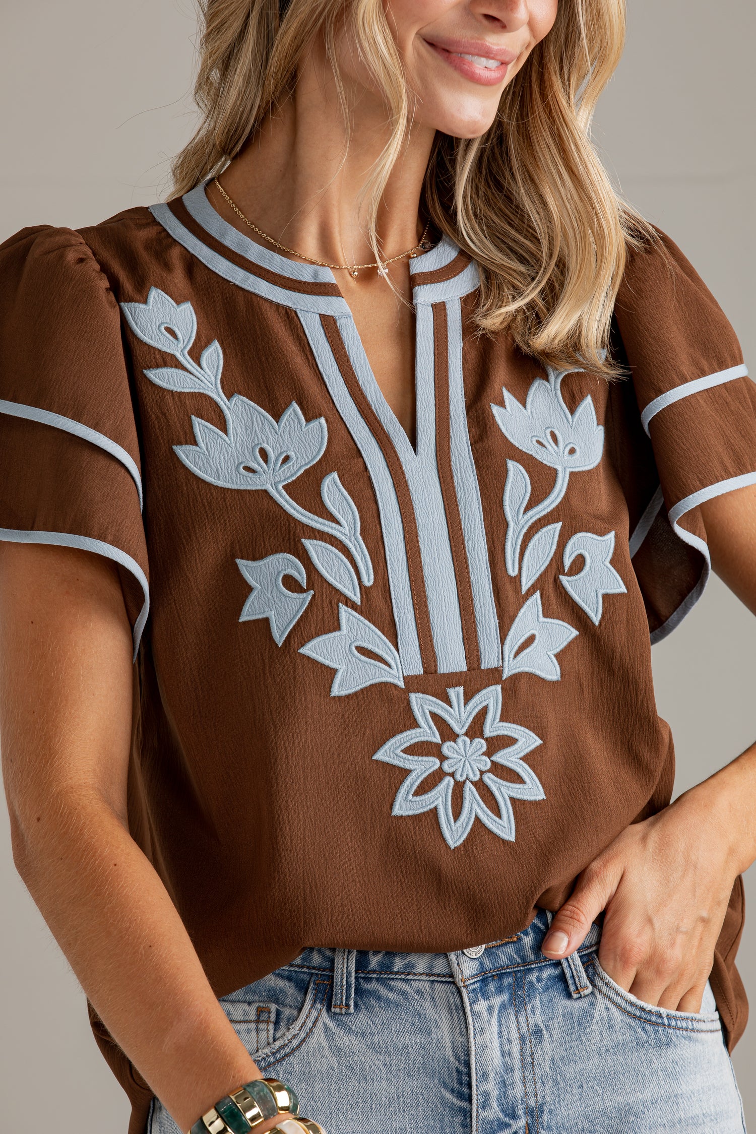 A woman wearing a brown short-sleeve blouse with white floral embroidery on the front and light gray piping along the neckline and sleeve cuffs, paired with light-wash blue jeans.