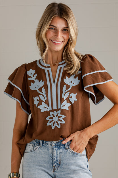 A woman wearing a brown short-sleeve blouse with white floral embroidery and light blue jeans, standing against a light gray paneled wall.