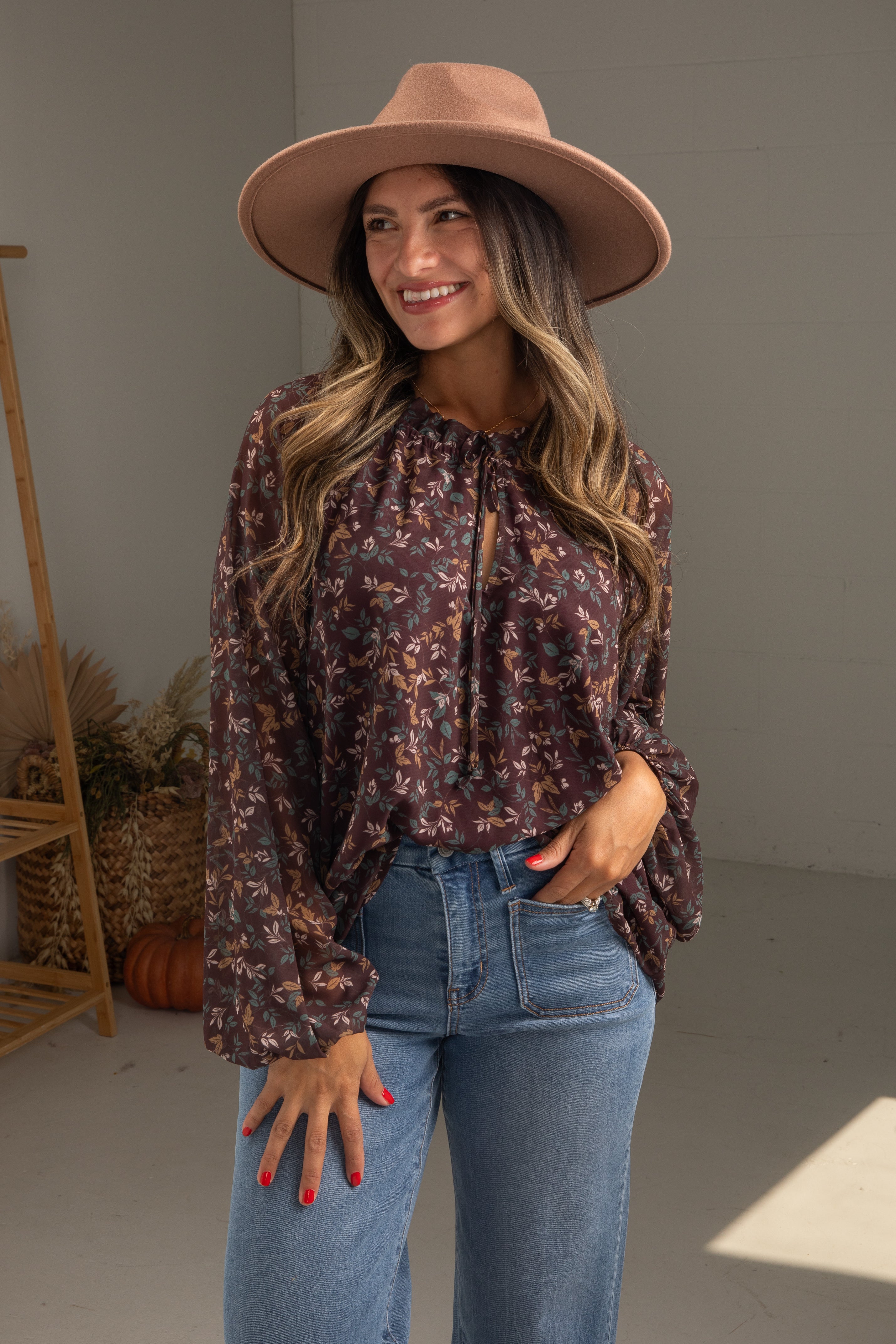 Woman wearing a floral blouse, wide-brimmed hat, and jeans indoors.