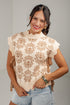 A woman wearing a sleeveless cream knit sweater vest with a brown floral pattern and ruffled edges, paired with light blue denim shorts, against a neutral gray studio background.