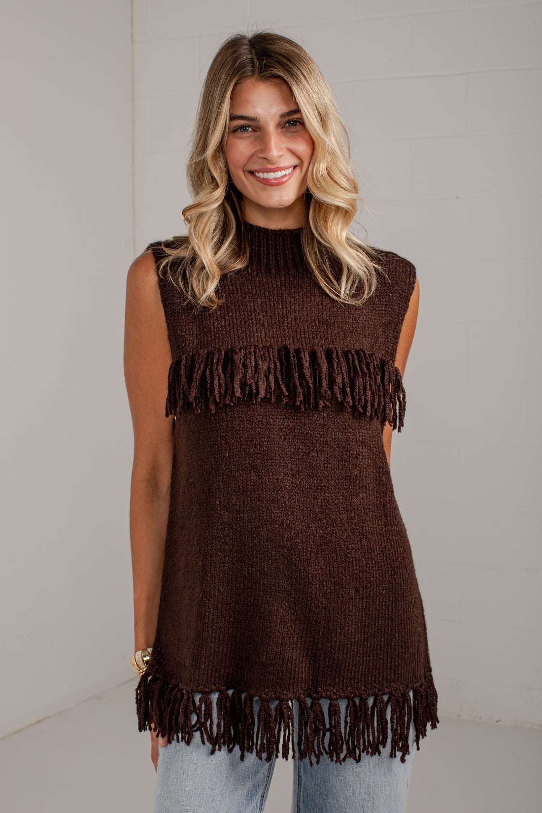 A woman wearing a sleeveless dark brown knit tunic top with fringe trim at the chest and hem, paired with light blue jeans, standing against a light gray wall.