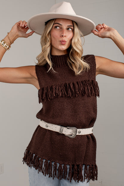 A woman wearing a sleeveless brown knit top with fringe trim, a white belt, and a wide-brimmed cream hat, posing against a light gray wall.