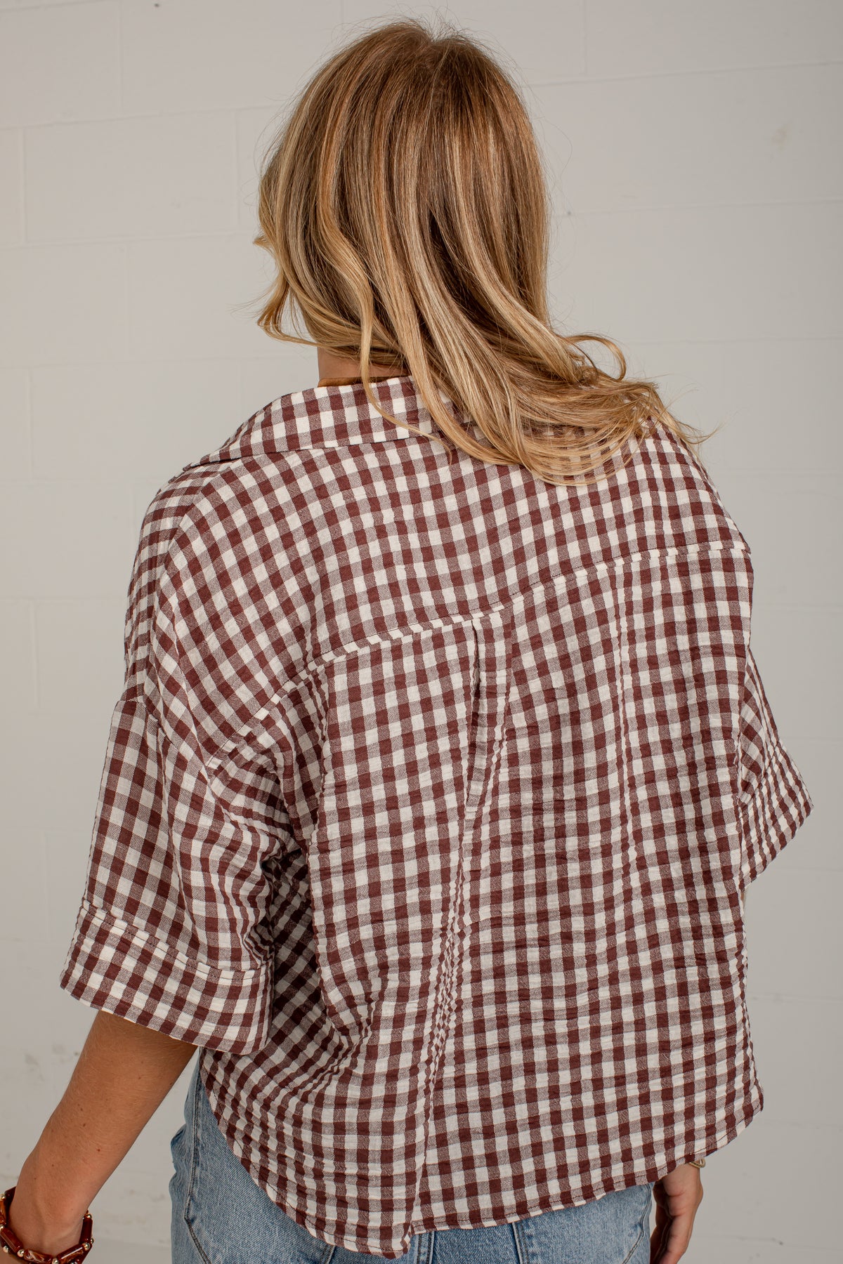 A woman viewed from behind wearing a brown and white gingham check shirt with rolled-up sleeves and light blue jeans, set against a plain off-white wall.