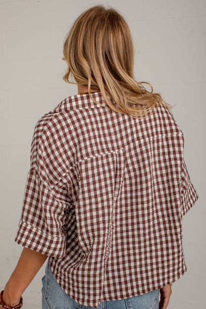 A woman viewed from behind wearing a brown and white gingham check shirt with rolled-up sleeves and light blue jeans, set against a plain off-white wall.