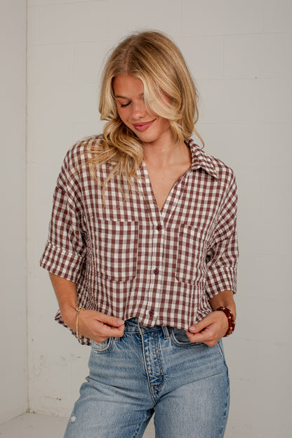 A woman wearing a brown and white gingham plaid button-down shirt with three-quarter sleeves and blue jeans, standing against a white wall.