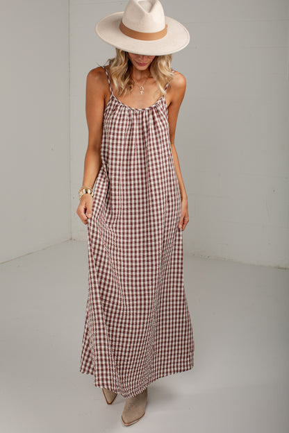 A woman modeling a brown and beige gingham maxi dress with spaghetti straps, styled with a wide-brimmed cream hat and beige ankle boots.
