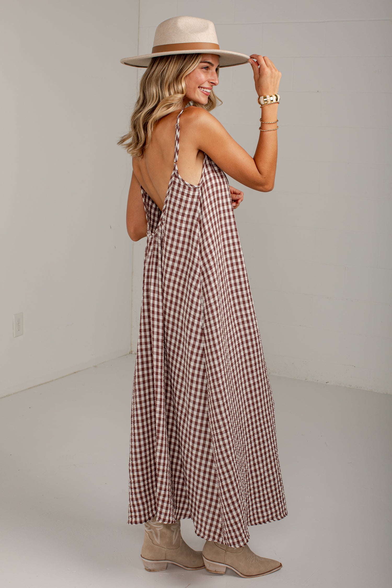 A woman wearing a brown and white gingham maxi dress with a backless open design, styled with a beige wide-brim hat and beige ankle boots, posing in a minimalist studio setting.
