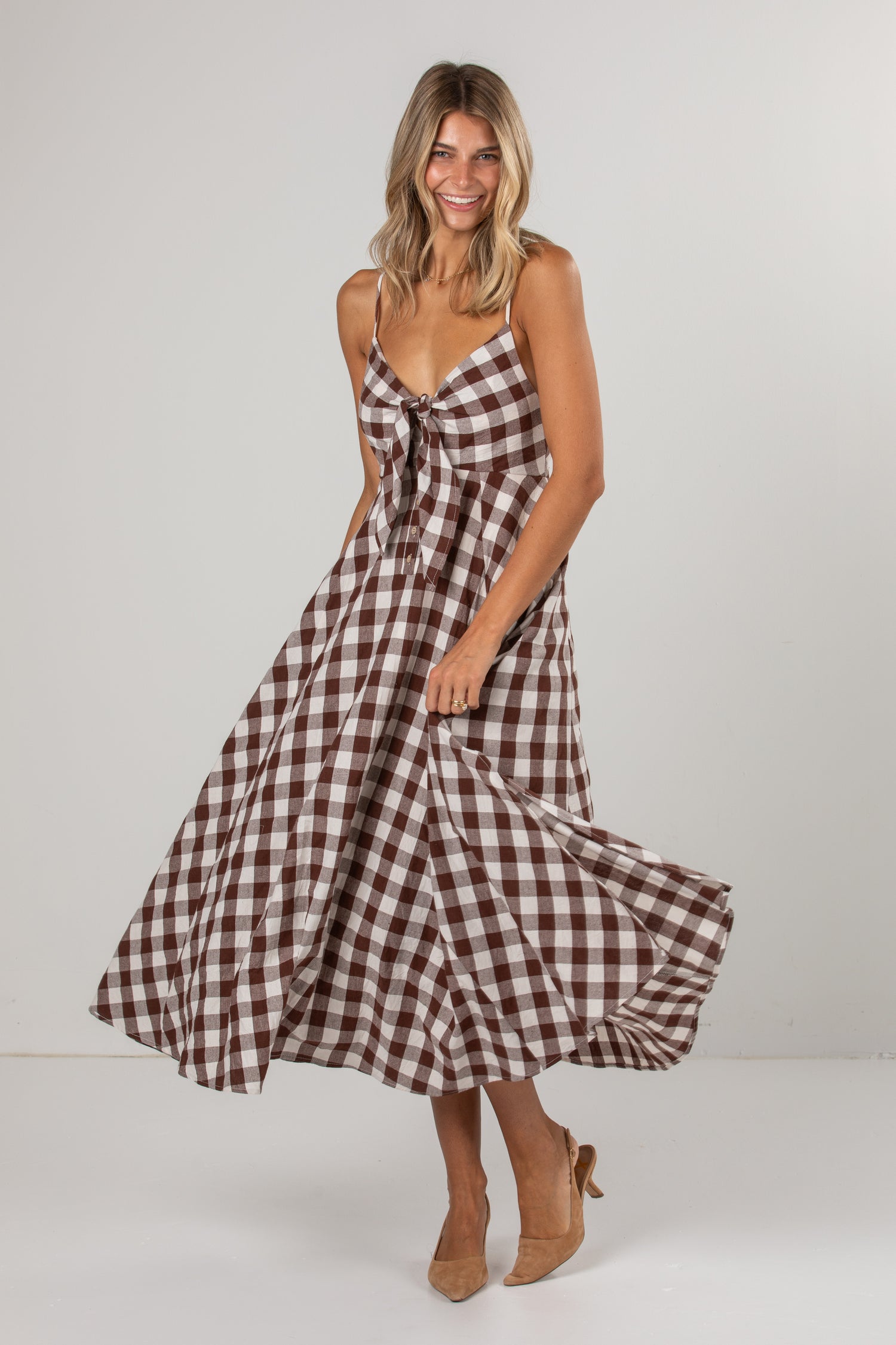 A woman modeling a brown and white gingham midi dress with spaghetti straps and a tie detail at the bust, standing against a light gray studio background.