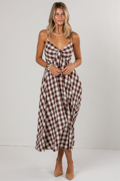 A woman modeling a brown and white gingham midi dress with spaghetti straps and a gathered waist, paired with tan high heels against a plain white studio background.