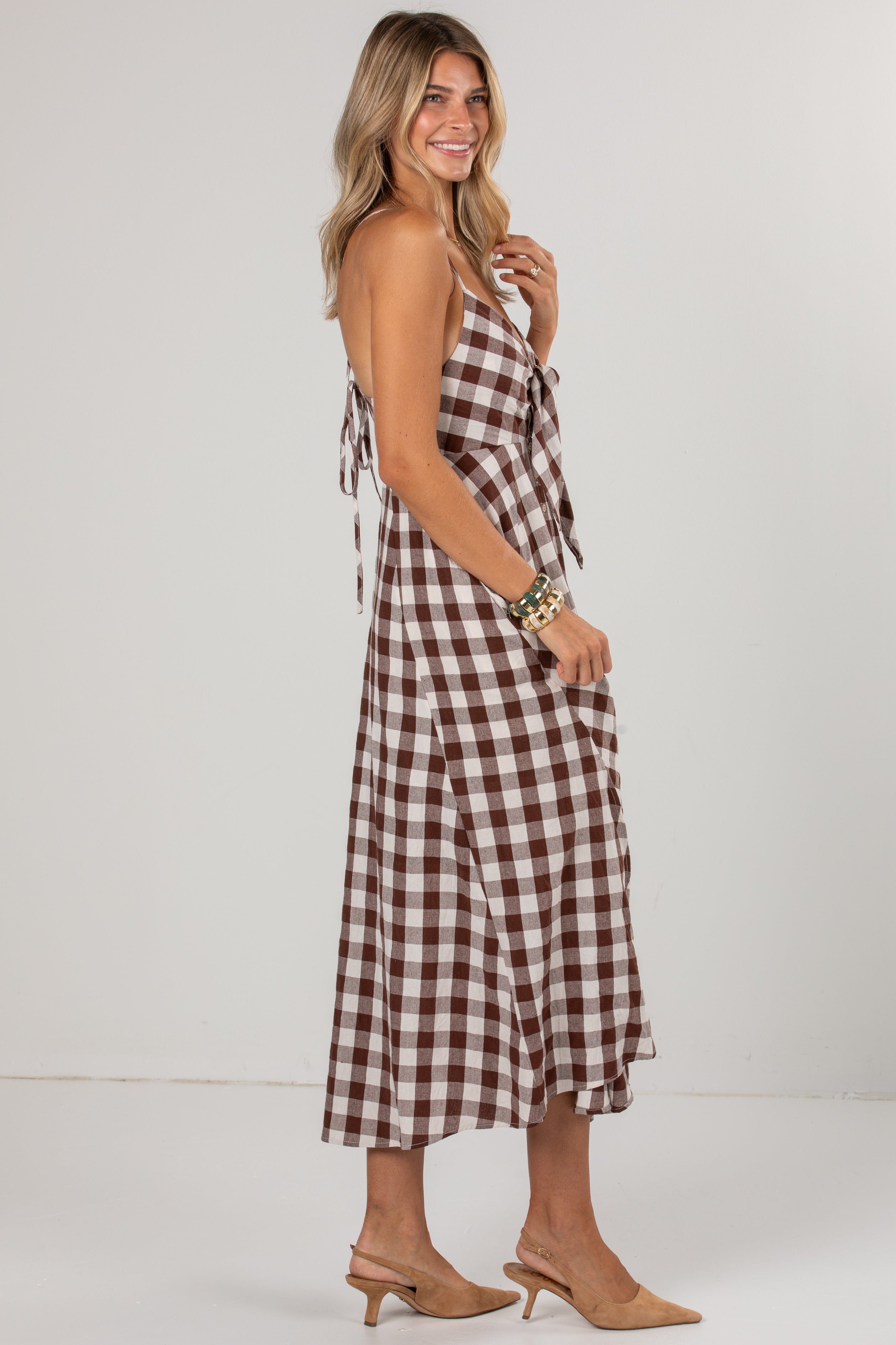 A woman modeling a brown and white gingham wrap midi dress with tie straps, paired with beige slingback heels, against a plain white studio background.