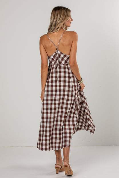 A woman modeling a brown and white gingham midi dress from the back, featuring thin adjustable tie-shoulder straps and a flowing skirt, set against a plain light gray studio background.